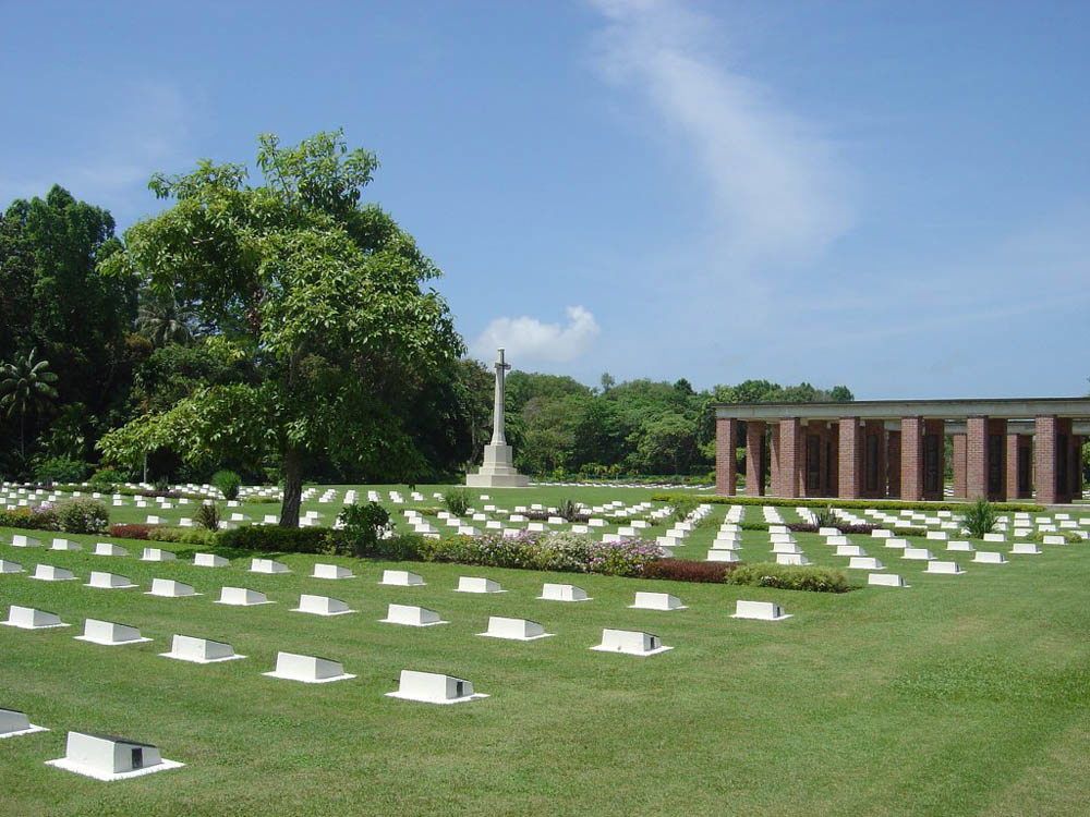 Headstone markers stretching over a green lawn at Labuan War Cemetery. The red brick, rectangular structure know as the Labuan Memorial, is visible in the background.