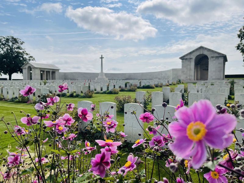 Scene showing headstones, cross of sacrifice, and rear portico at Serre Road No.2 Cemetery. Bright pink and yellow flowers are visible in the foreground.