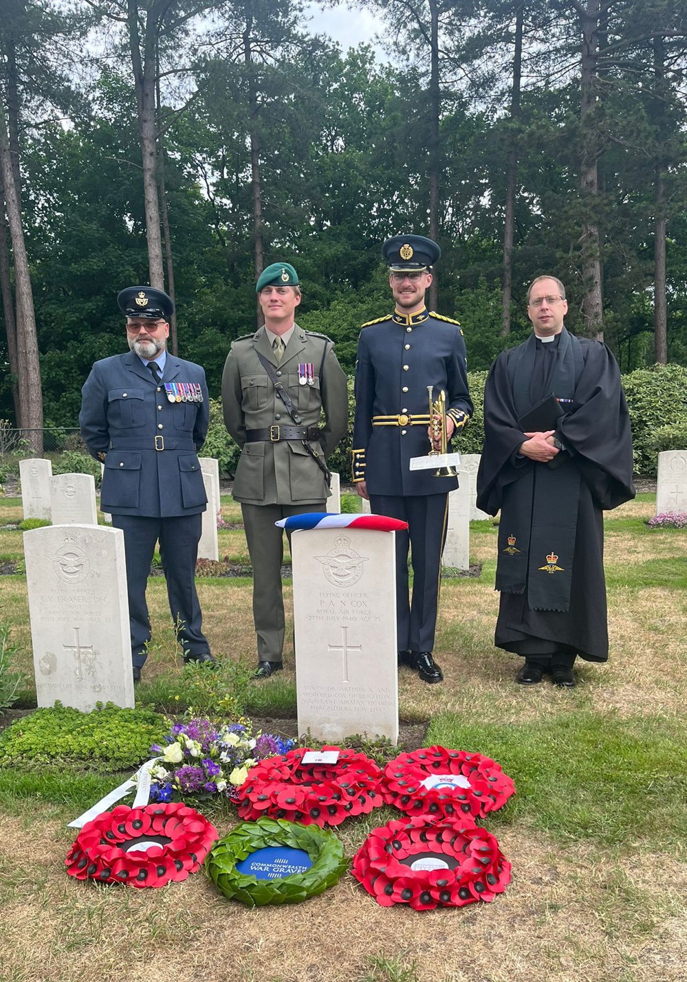 Military officers and a priest stand around the grave of a WW2 flying officer. A union jack has been laid on the pilot's CWGC headstone and several red wreaths have been laid before his grave.