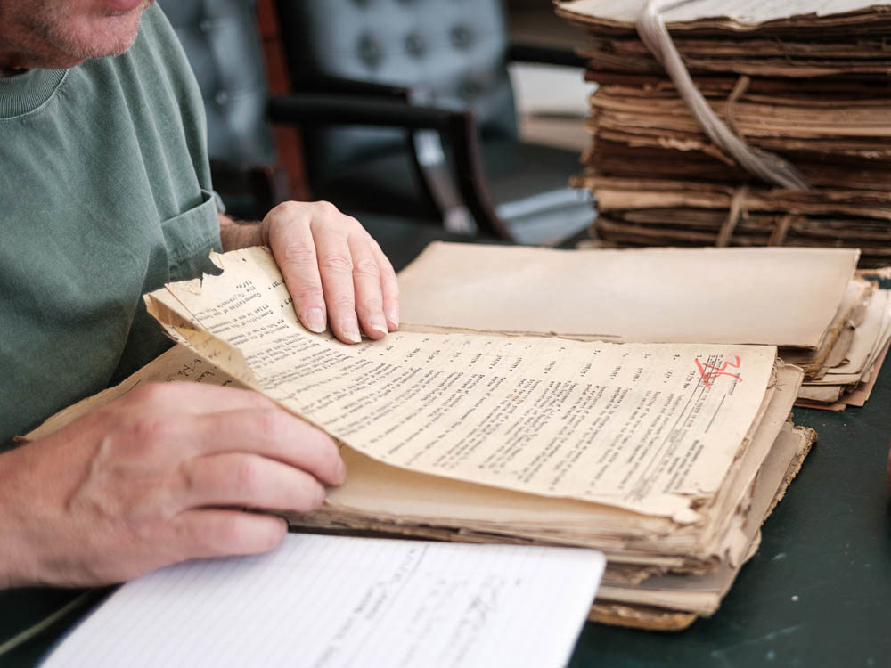 A man reviewing old, yellowed documents in Kenyan archives. A pile of old, twine-bound pile of records sits on the desk next to the investigator.