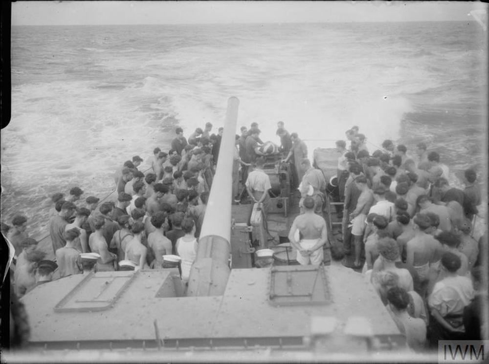 Crewmen of HMS Qullion prepare to commit a Union Jack-draped coffin into the Pacific Ocean. The crew are gathered beneath one of the ships rear facing naval guns.