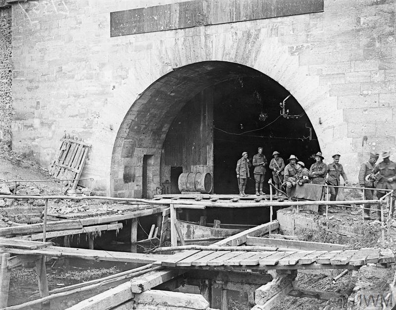 Australian and American soldiers meeting beneath a huge stone tunnel at the St Quentin Canal. The canal chennel feeds into the tunnel, which is arched with large wooden gates.