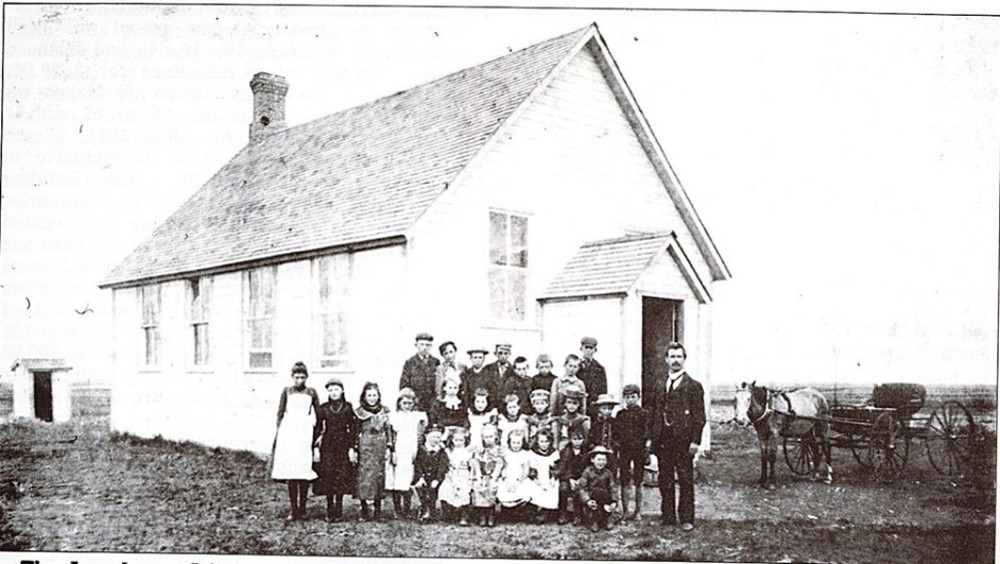 Canadian schoolchildren posing outside their small white brick school building some time before the turn of the 20th century.