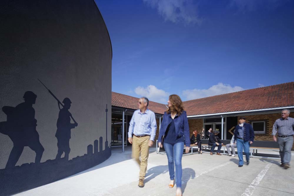 Visitors walk through the central coutryard at the CWGC Visitor Centre in Beaurains, France. The sky is a bright blue dappled with whisps of cloud. Around the courtyard are flat, red roofed buildings and a round curtain wall. The wall is silhouetted with images of headstones and First World War era soldiers.