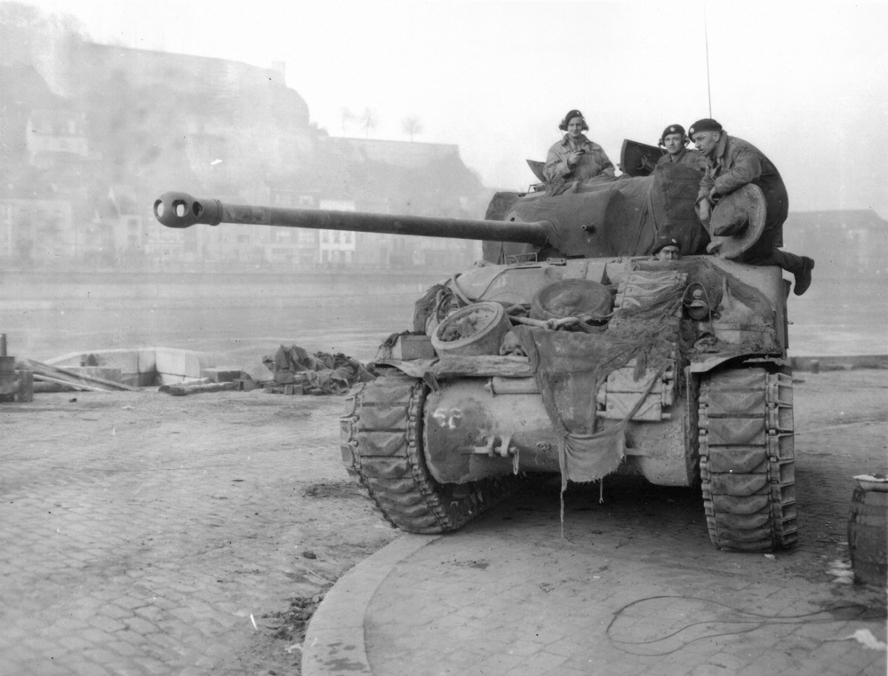 A Sherman Firefly tank with near the River Meuse at Namur, Belgium. Two tankers are visible in the turret while another sits atop the tank. The driver's head is visible poking through his vision hatch in the hull.