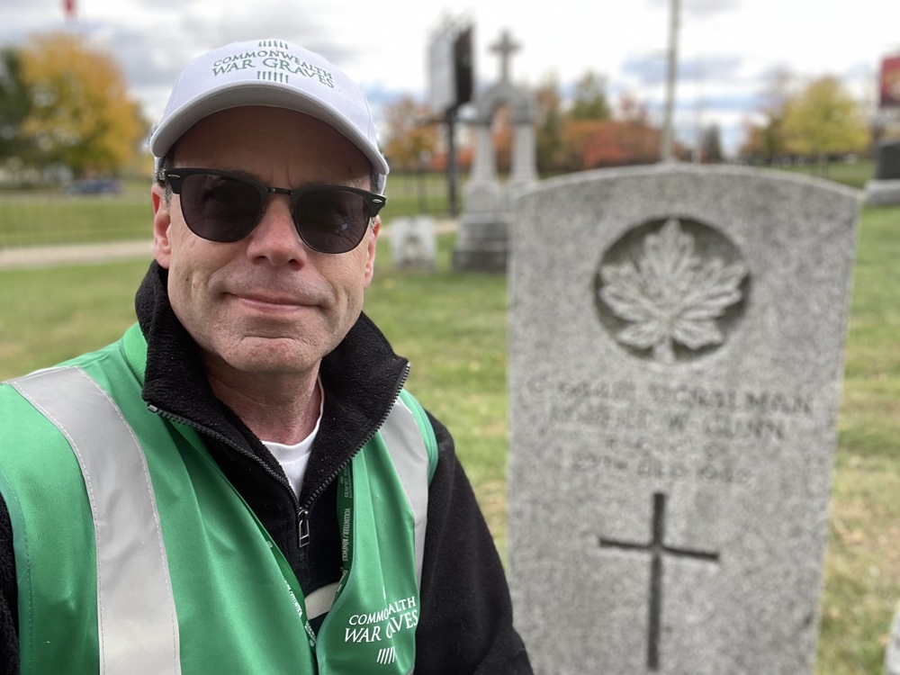 Stuart, a Canadian volunteer, poses next to a dark grey stone CWGC headstone within a Canadian cemetery. Stuart is wearing a green hi-vis vest with the CWGC logo and white CWGC cap.