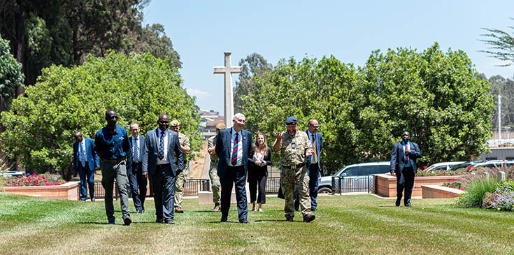Group of dignitaries and military figures walking through a Kenyan CWGC cemetery with the Cross of Sacrifice visible in the background.