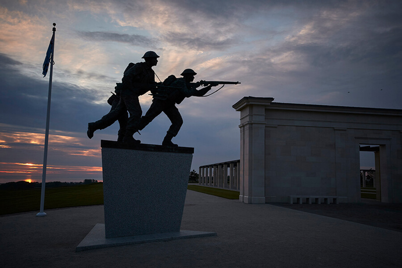 The British Normandy Memorial at dusk with the bronze statue of advancing British infantrymen in the foreground.