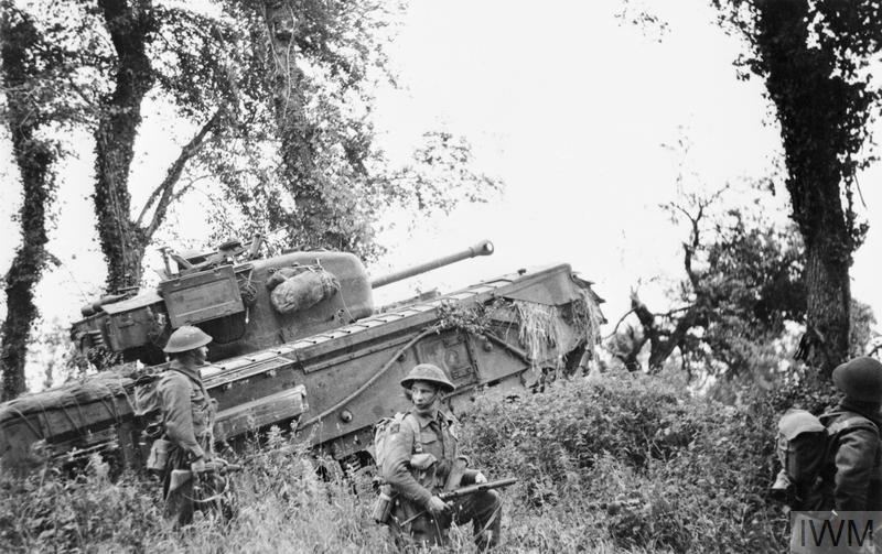 A Churchill Tank advances through woods while supported by Scottish infantry.