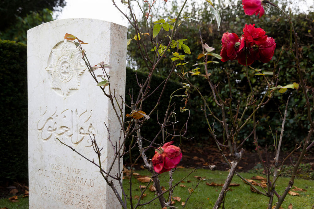 Headstone of Gul Muhammad, Indian Army Service Corps, showing inscription in arabic text with the four-pointed star cap badge of his regiment. A slighlty bare rose plant spiders its skeletal branches over the grave. Several red flowers still bloom but the plant is dying as autumn approaches.