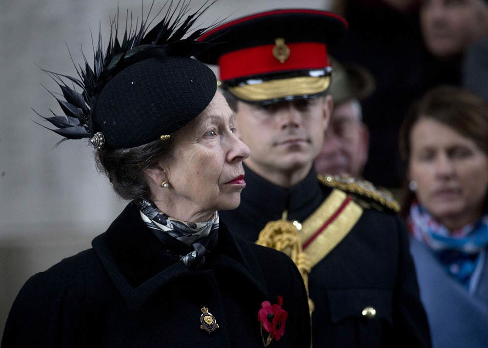 Here Royal Highness THe Princess Royal weaing a feathered black hat and long black coat for Armistice Day ceremonies at the Menin Gate Memorial. A soldier in a red, gold brimmed cap and dark jacket with red and gold sash stands behind the Princess.