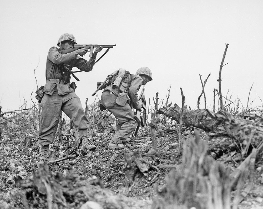 US marines pick their way through smashed, overchurned ground on Okinawa, Stumps and logs block their path. The second marine is taking aim with his Tommy gun while his friend takes a low running stance to avoid enemy fire.