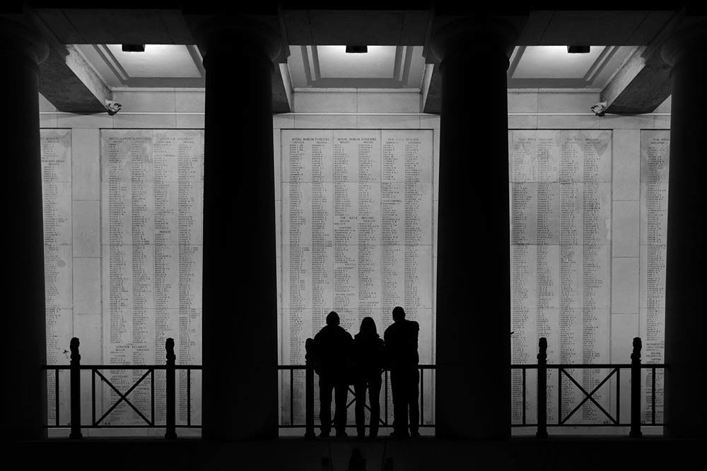 Three figures observing the interior name panels of the Menin Gate Memorial silhouetted black against the panel's grey.