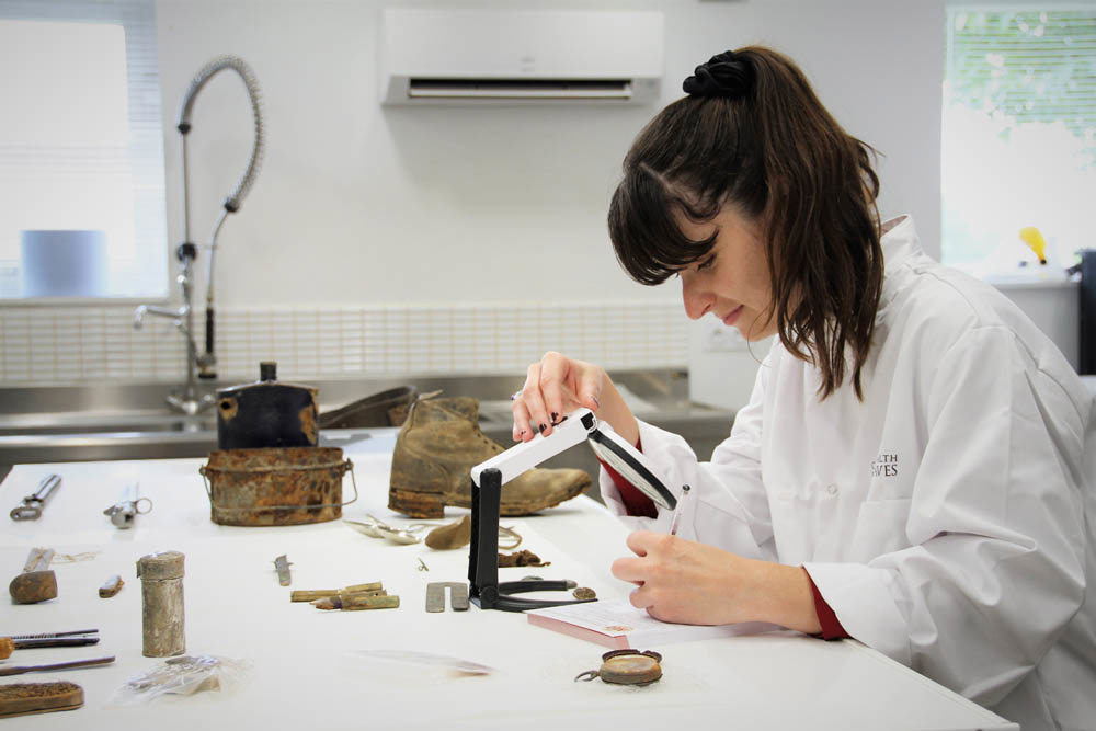 Women in a white lab coat inspects WW1-era artefacts with a large magnifying glass.