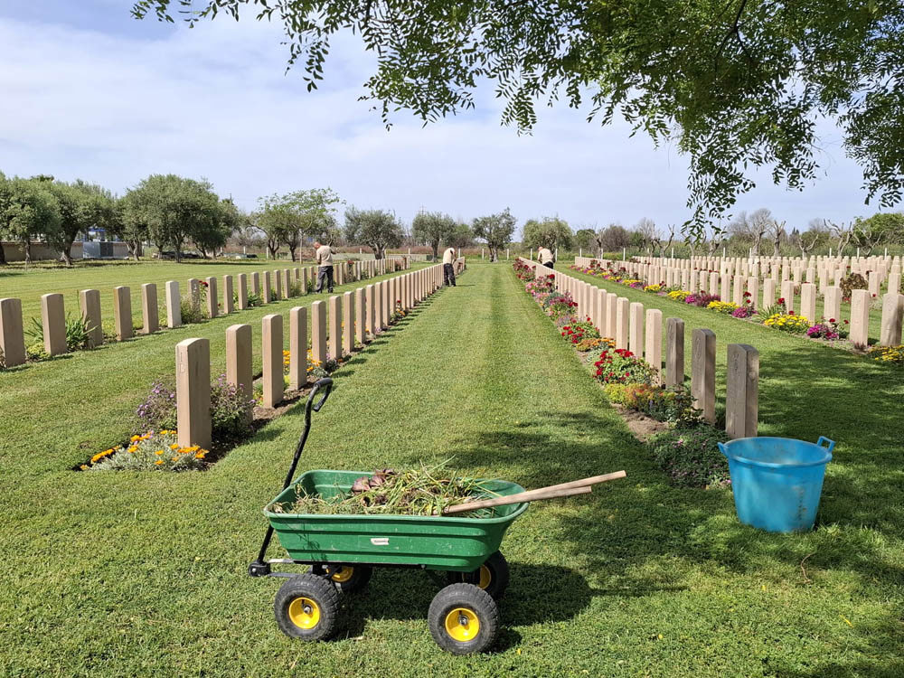 A wheelbarrow full of mulch and dirt sits at the end of a headstone row in Catania war cemetery. In the background, you can see the gardeners working on plant borders and the green lawn.