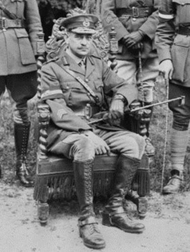 General John Monash sits in his military uniform on a cap chair somewhere on the Western Front. He is flanked by other officers but their faces are obscured.