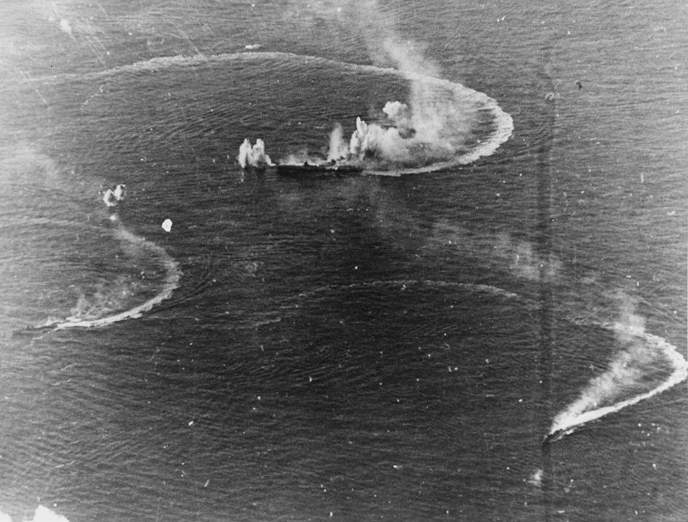 An aerial view of Japanese Navy vessesl under attack by American aircraft at the Battle of the Philippine Sea. Huge splashes can be seen where US bombs have hit the water. White smoke erupts from the ship in the top middle of the image as bombs strike its superstructure.