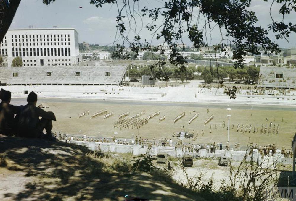 A long range view of British and Allied troops arrayed in lines ahead of an atheltics event in Rome, 1944. Two British soldiers can be seen watching proceedings underneath a tree in the foreground.