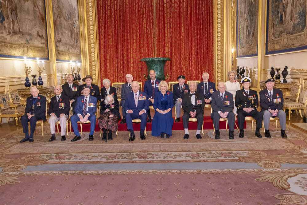 King Charles III, Queen Camilla, veterans, and members of the Armed services pose in front of a giant green vase in a fancy ballroom in Windsor Castle.