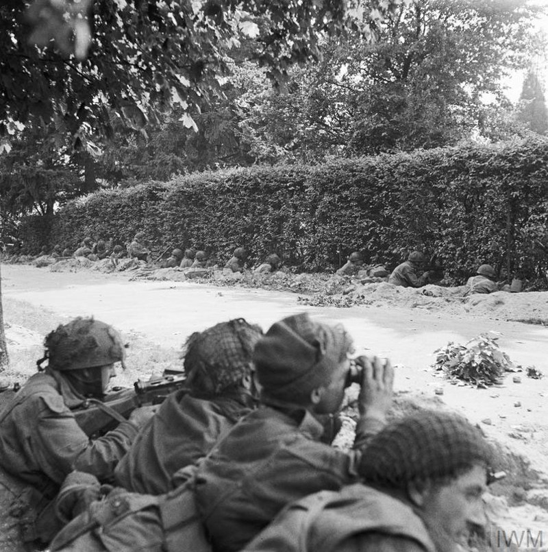 British paratroopers taking cover in a trench. A further line of soldiers is hiding behind a thick, tall hedgerow in front of the trench.