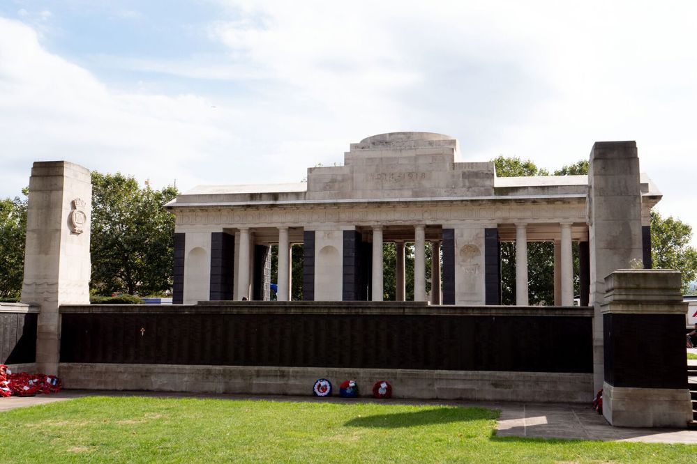 The Tower Hill Memorial as seen from the Second World War garden showing a memorial wall featuring bronze name panels flanked with two small towers ahead of the original First World War memorial building. Some wreaths have been layed against the memorial wall.