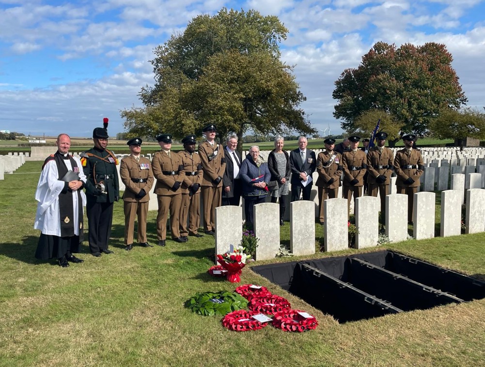 A row of British soldiers, a priest, and family members pose next to open war graves, ready for burial. A row of CWGC headstones sits in front of the burial plots, Red poppy wreaths are visible to the left.