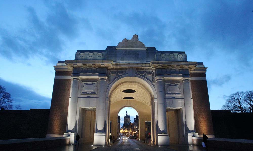 View of the Menin Gate in the evening, illuminated from within. Through the triumphal arch, Ypres cloth hall, streetlights and a car can be seen. The sky, is still a pale blue but dappled with clouds of various dark blue hues.