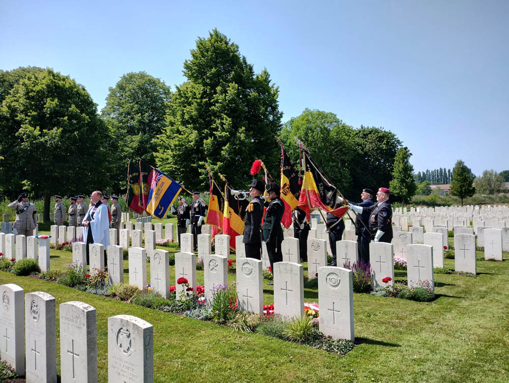 Standards are lowered as the Last Post sounds in a CWGC cemetery during a rededication ceremony. Uniformed military members hold a mixture of Belgian and British military flags. A vicar is giving a reading.