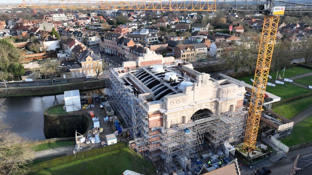 Menin Gate under restoration works in 2024. The front of the entrance hall is covered in scaffolding. A large yellow and black crane stretches high over the memorial. Various pieces of building equipment are visible in a storage yard to the the left of the main hall. Ieper town centre is visible behind the memorial.