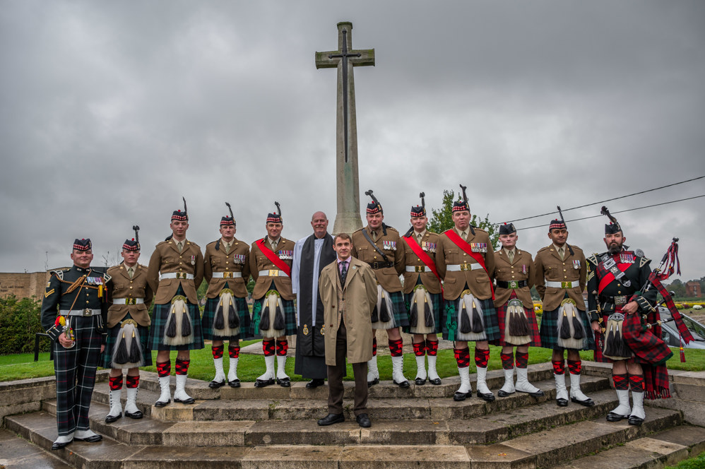 Man in brown overcoat poses with priest and Scottish soldiers in kilts around a CWGC Cross of Sacrifice