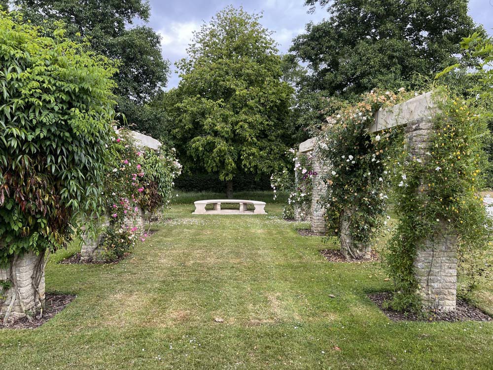 A lawn with white daisies. It is flanked by stone structures covered in plant life with a stone bench visible at the end of the lawn.