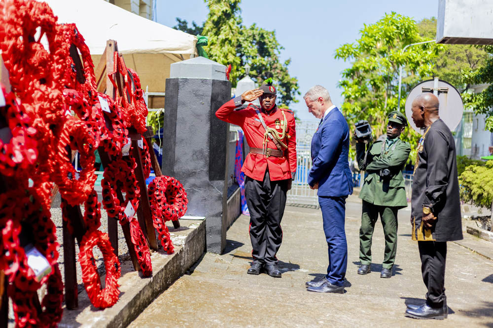 A Sierra Leonean soldier in red field dress salutes next to a wall of wreaths laid at the Freetown Memorial while other figures bow their heads in respect.