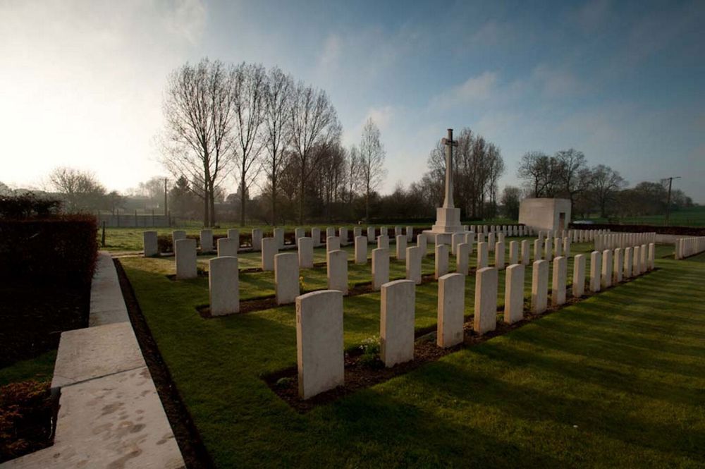 View of Forceville Communal Cemetery & Extension at dusk. Clearly visible is a section of the stone boundary wall, rows oof CWGC headstones, the Cross of Sacrifice, and the squat, square shelter building tucked in the far right corner.