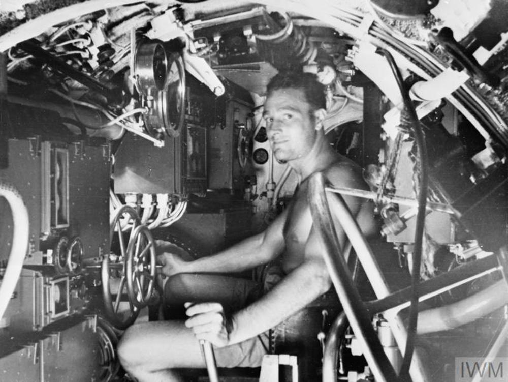 View of a shirtless Submariner operating the diving station of his WW2 Midget submarine.The interior of the sub is very cramped, full of dials, wheels, pipes, levers, and assorted technologies.