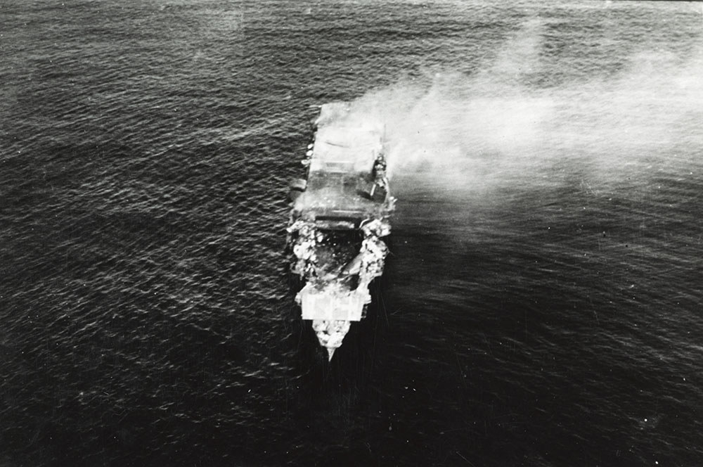 The damaged Japanese Aircraft carrier Hiryu billows white smoke as she slowly sinks beneath the waves. A huge hole has been gouged in the flight deck by enemy naval gun fire.
