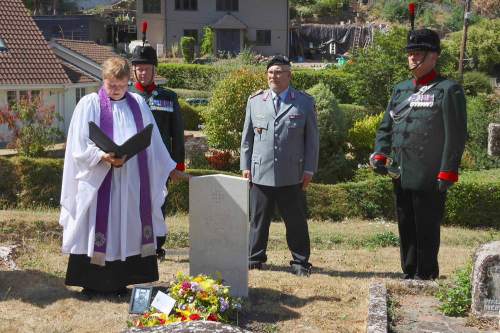 Priest touching a German airman's headstone while Bundeswehr officials look on behind.