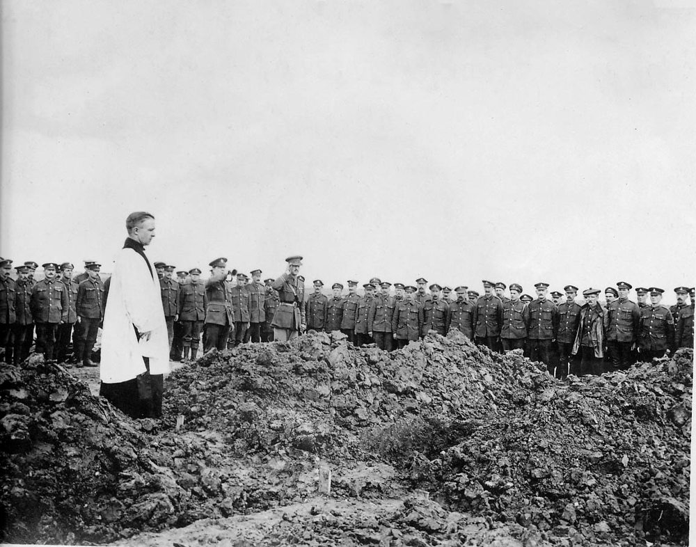 An army Chaplain leads a sombre ceremony at a mass grave. Great clods and mounds of earth covere the burial site. In the background, soldiers stand to attention while a bugler plays and an officer salutes.