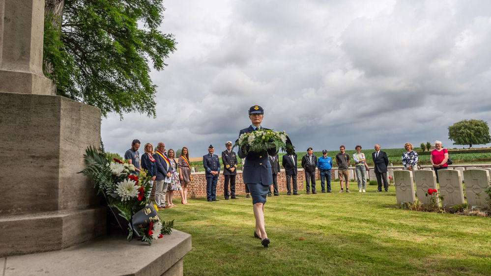 A New Zealand service woman in dark grey military uniform appraches a CWGC Cross of Sacrifice to lay a green wreath for remembrance.