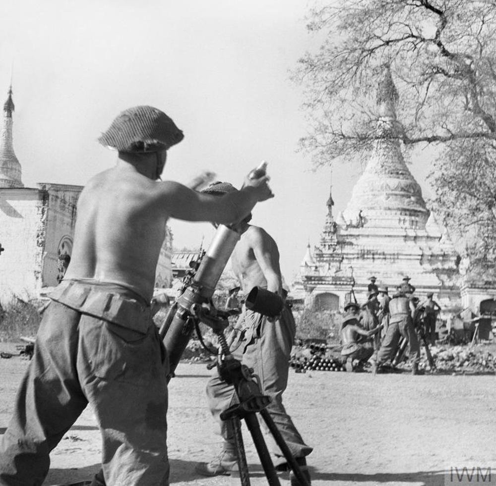 A shirtless British mortar crew loads their weapon in the streets of Meiktila, Burma, circa February 1945. Another mortar crew is visible in the background as is a large golden pagoda.