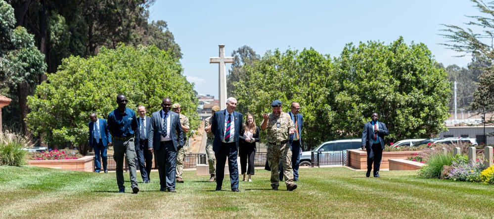 Group of dignitaries and military figures walking through a Kenyan CWGC cemetery with the Cross of Sacrifice visible in the background.