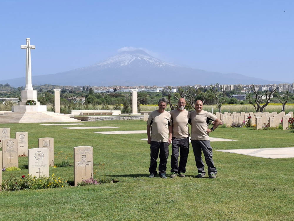 Gardeners at Catania War Cermetery pose cheerfully for the camera. The Cross of Sacrfice is visible in the left while Mount Etna looms large in the background.