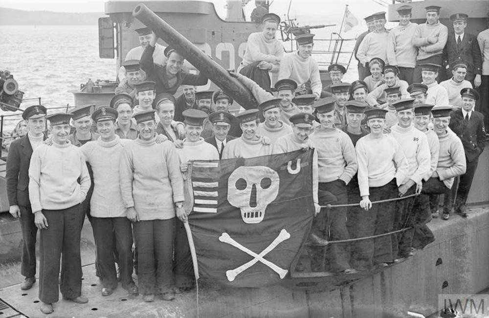 The crew of HMS Satyr posing on the deck of their submarine holding a up a Jolly Roger flag.