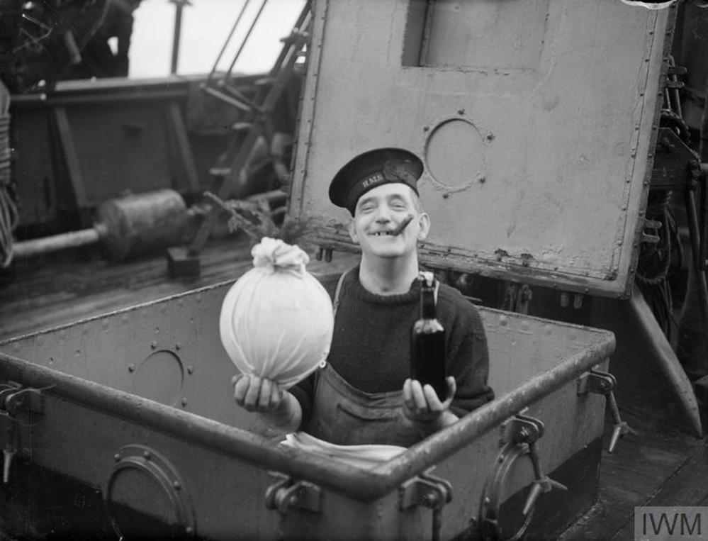 A British Royal Navy sailor holding a Christmas pudding and a bottle of beer in the hatch of his warship.