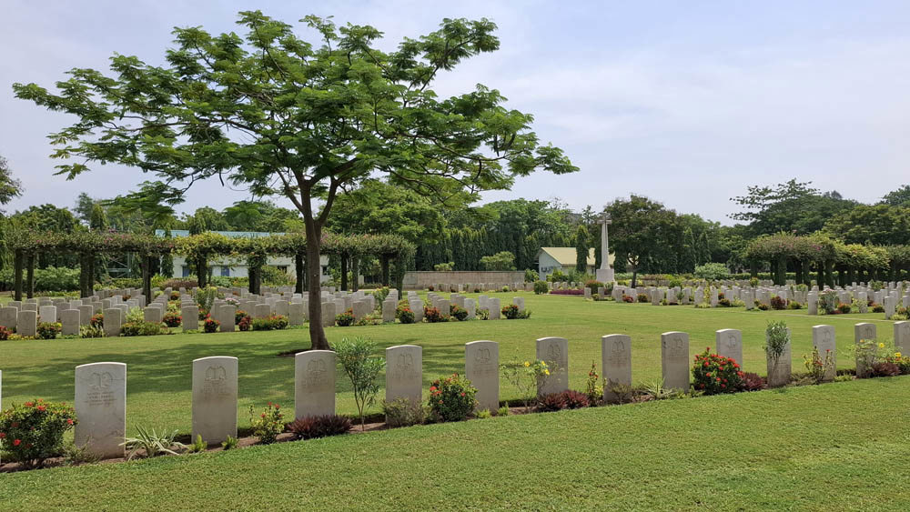 View of Madras War Cemetery showing a row of white CWGC headstones on a green lawn. A tall tree with broad green leaves is visible behind the row. You can also see a vine-covered shelter building in the background, opposite the CWGC Cross of Sacrifice.