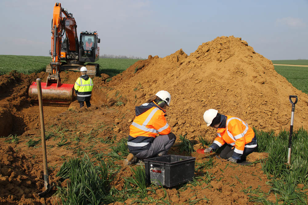 Men in hi vis vests digging in a field next to a large mound of dirt. A digger is carving out a trench in the background.