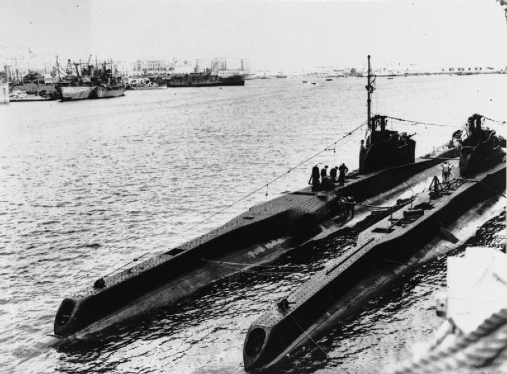 HMS Turbulent, left, docked next to another T Class submarine in a British harbour. Various warships of different sizes can be seen in the background.