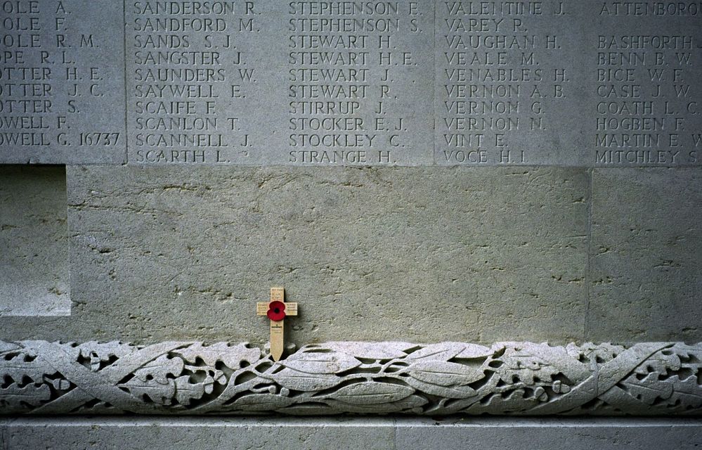 Intricate stonework showing leaves and vines carved into a border at the Thiepval Memorial. A small wooden cross adorned with a poppy has been placed at the base of a name panel.