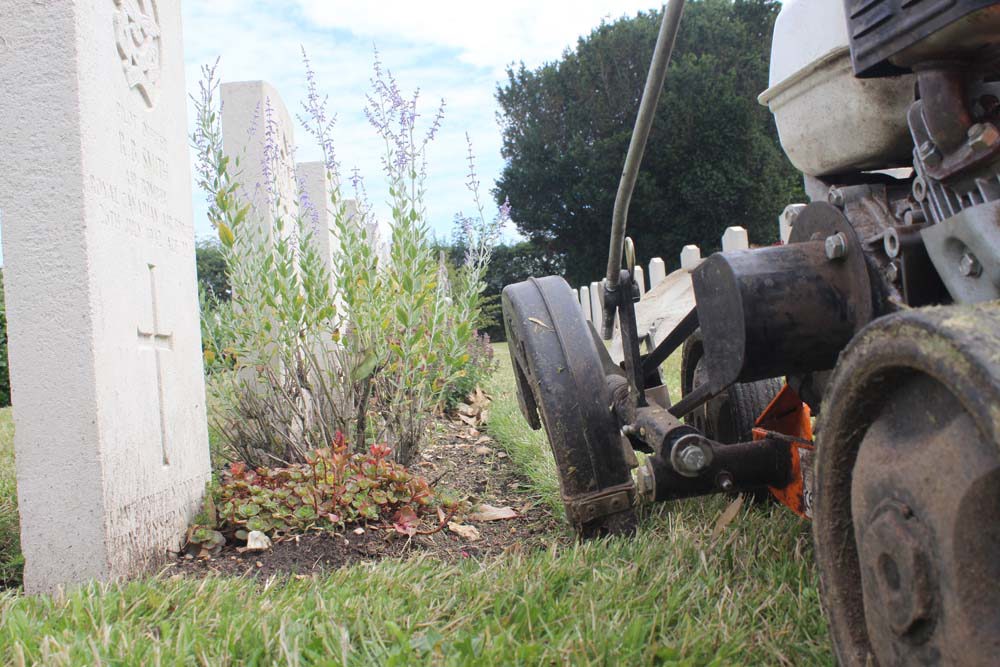 Close up of a push lawnmower mowing grass next to a CWGC headstone border.