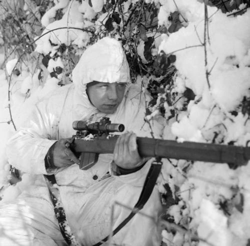 A British paratrooper sniper in white winter camouflage suit hiding amidst snow-covered trees in the Ardennes during the Battle of the Bulge.