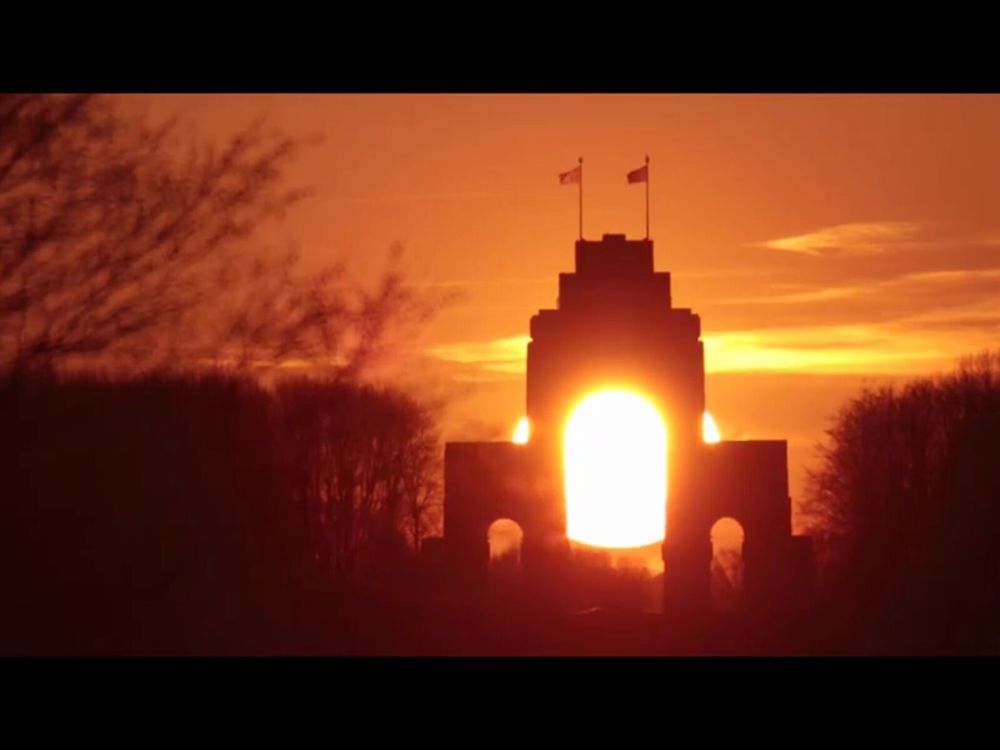 The sun setting behind the Thiepval Memorial. The low orange sun shines clearly through the central arch of the memorial, turning the entire seen a warm orange glow.